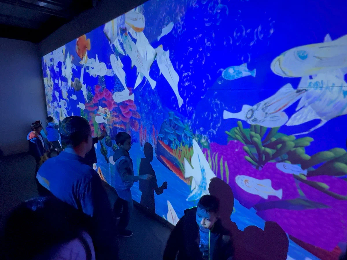 Kids and adults stand in an activity room with a lit wall full of animated fish.
