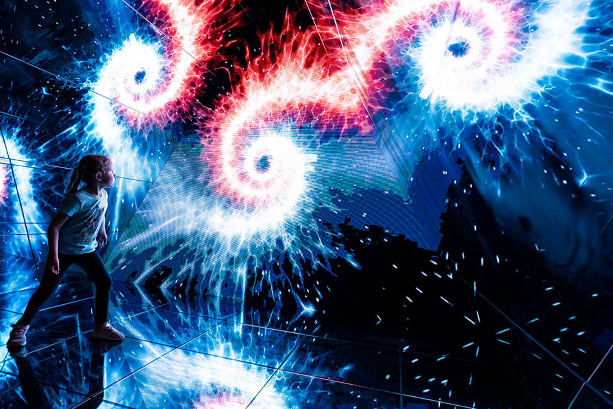 A young girl stares at a wall with a spectacular display of light bursts in a mirror room.