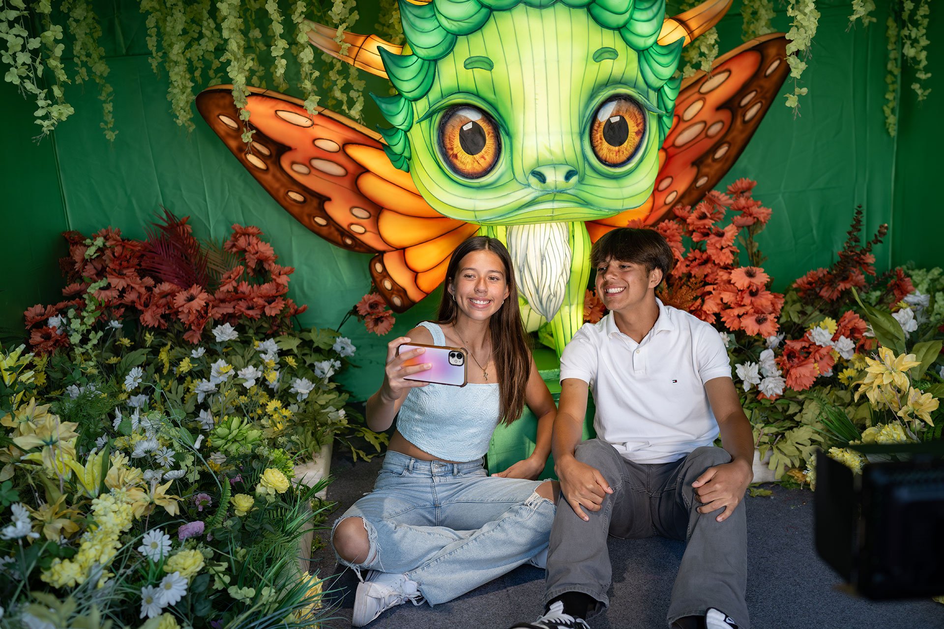 A teenage girl and boy take a selfie with a green magical creature behind them with flowers surrounding them.