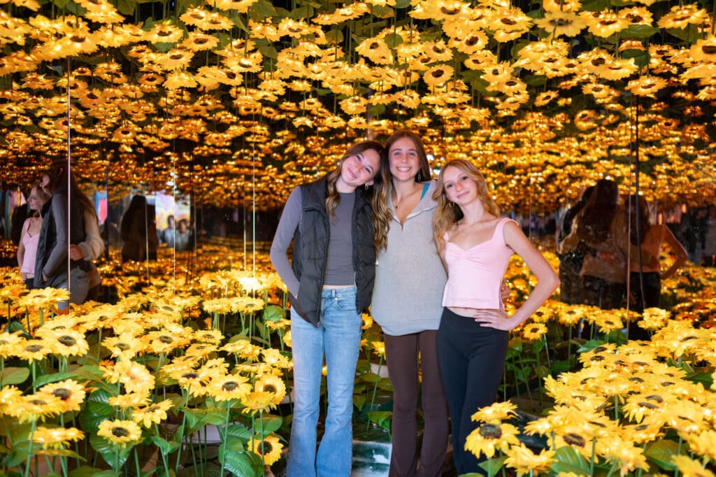 Three teenage girls pose for a photo in a room full of sunflowers.