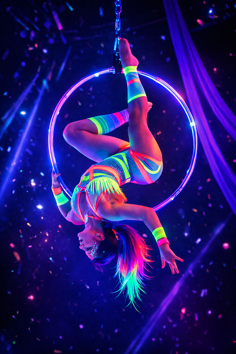 Aerial circus performer in neon outfit hangs upside down from a ring.