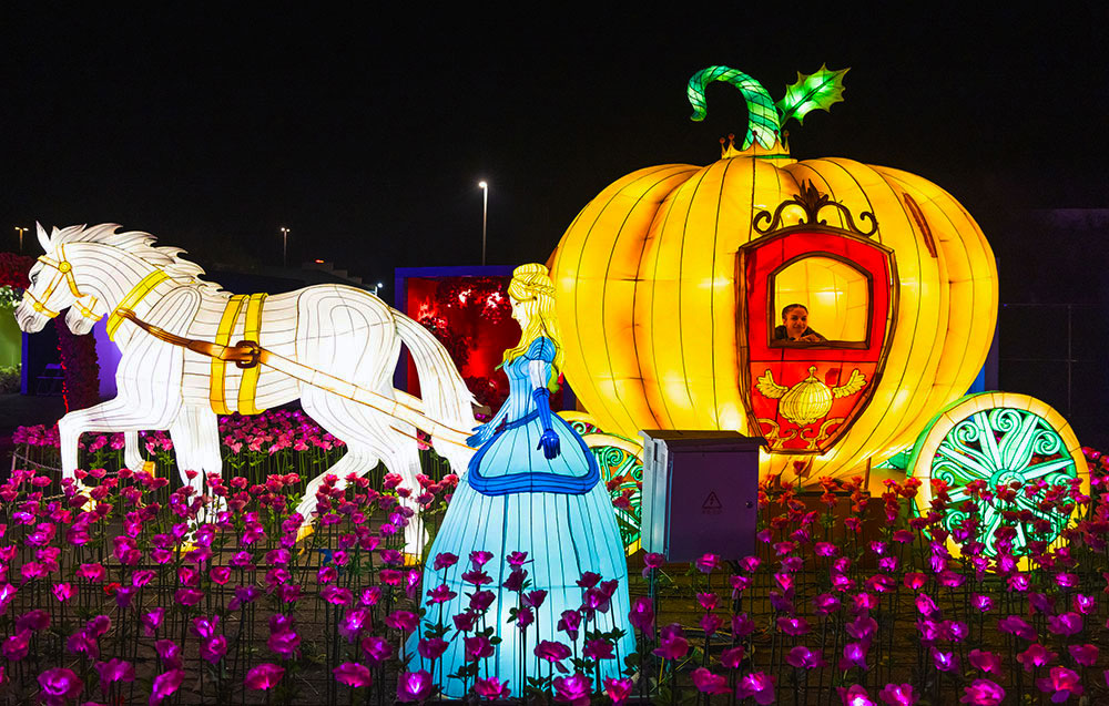A lighted structure of Cinderella standing in a field of lit flowers with a two horses pulling a pumpkin carriage behind her.