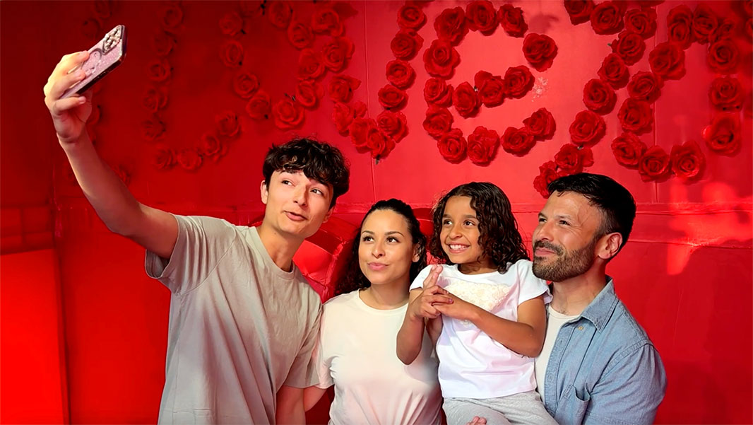A Hispanic family of four take a selfie photo in a red photo box with red roses.