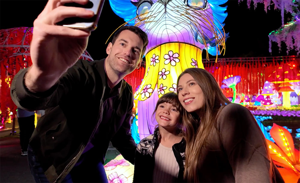 A caucasian family of three pose for a selfie with bright lit structures behind them.