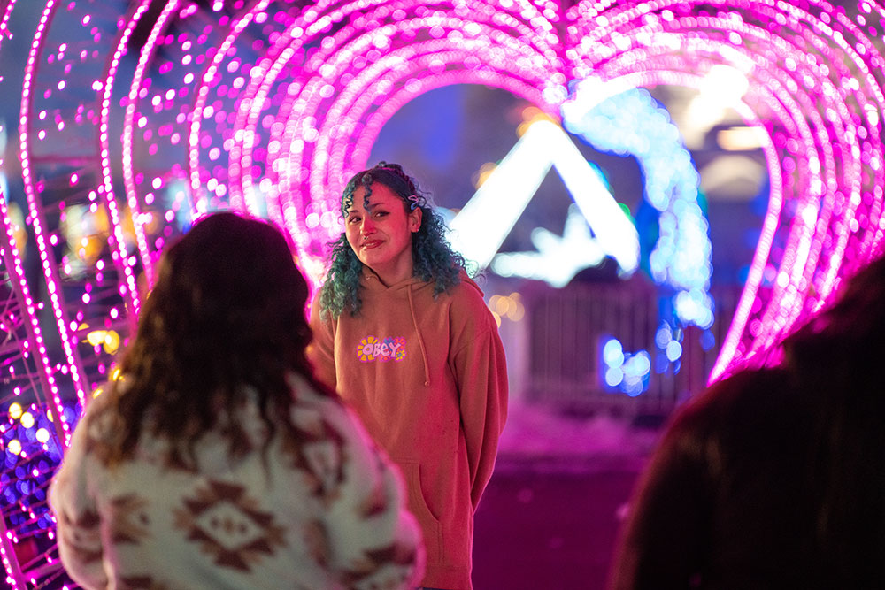 A young girl with blue hair poses for a photo with a pink lit heart shaped tunnel behind her.