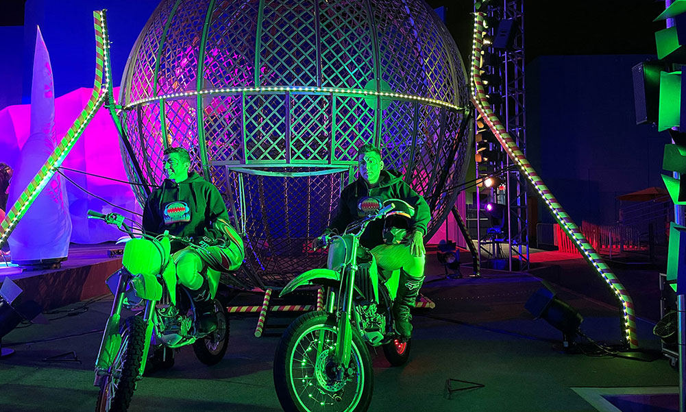 Two motorcycle riders sitting on their bikes in front of metal circular cage.