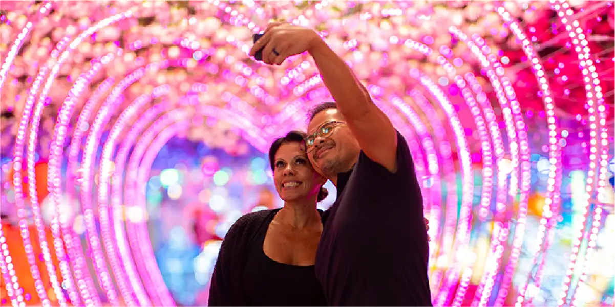 An adult male holds up his arm to take a selfie with an adult female while they stand in a pink lighted heart tunnel.
