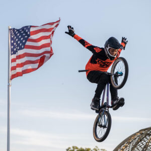 BMX Rider jumps through the air performing a trick with hands off handle bars. The American flag waves in the background