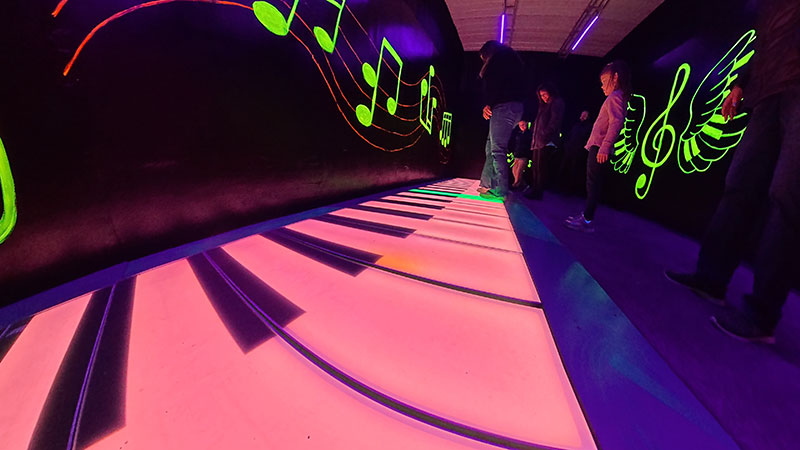 Imaginarium Let's Glow Crazy – In neon large piano room with neon music symbols on room, people play giant piano on floor.