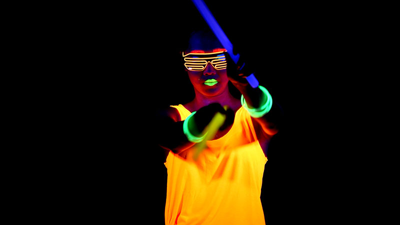 Imaginarium Let's Glow Crazy – Young woman with glowing drumsticks in neon shirt, glasses, and neon colored lips points at camera