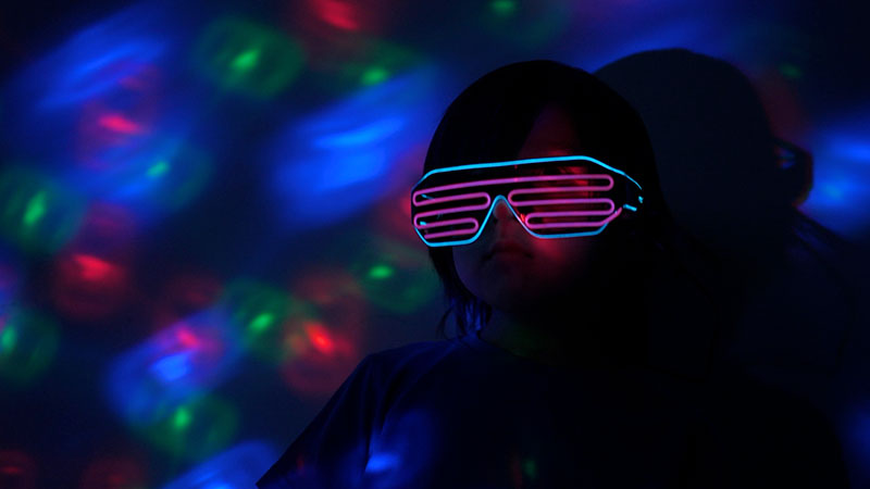Imaginarium Let's Glow Crazy – Boy with Neon sunglasses on in room with colored lights on wall