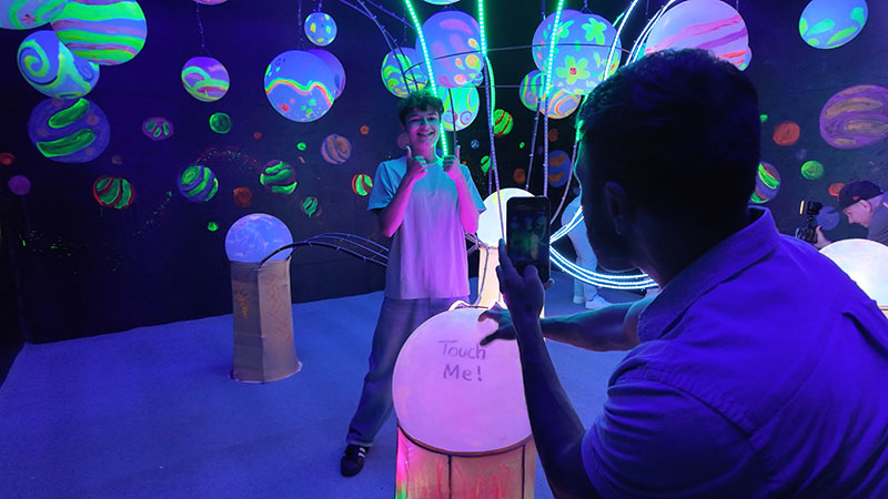 Imaginarium Let's Glow Crazy – In neon planet room father takes a photo of his son who gives two thumbs up to the camera