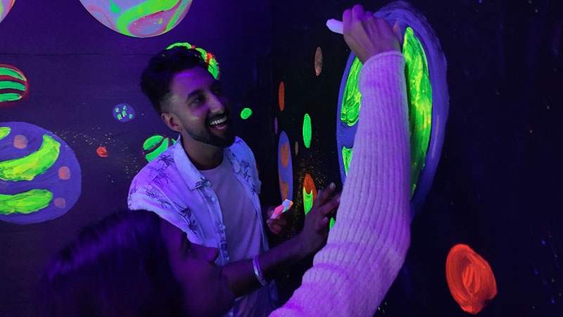 Imaginarium Let's Glow Crazy – In Neon Planet Room, young woman draws on the wall with a neon marker while young man smiles at drawing