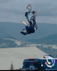 Imaginarium A person performs a mid-air backflip on a mountain bike above a van with mountainous scenery in the background.