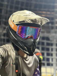Imaginarium Person wearing a motocross helmet with tinted goggles, dressed in a long-sleeve shirt, in front of a metallic grid background.